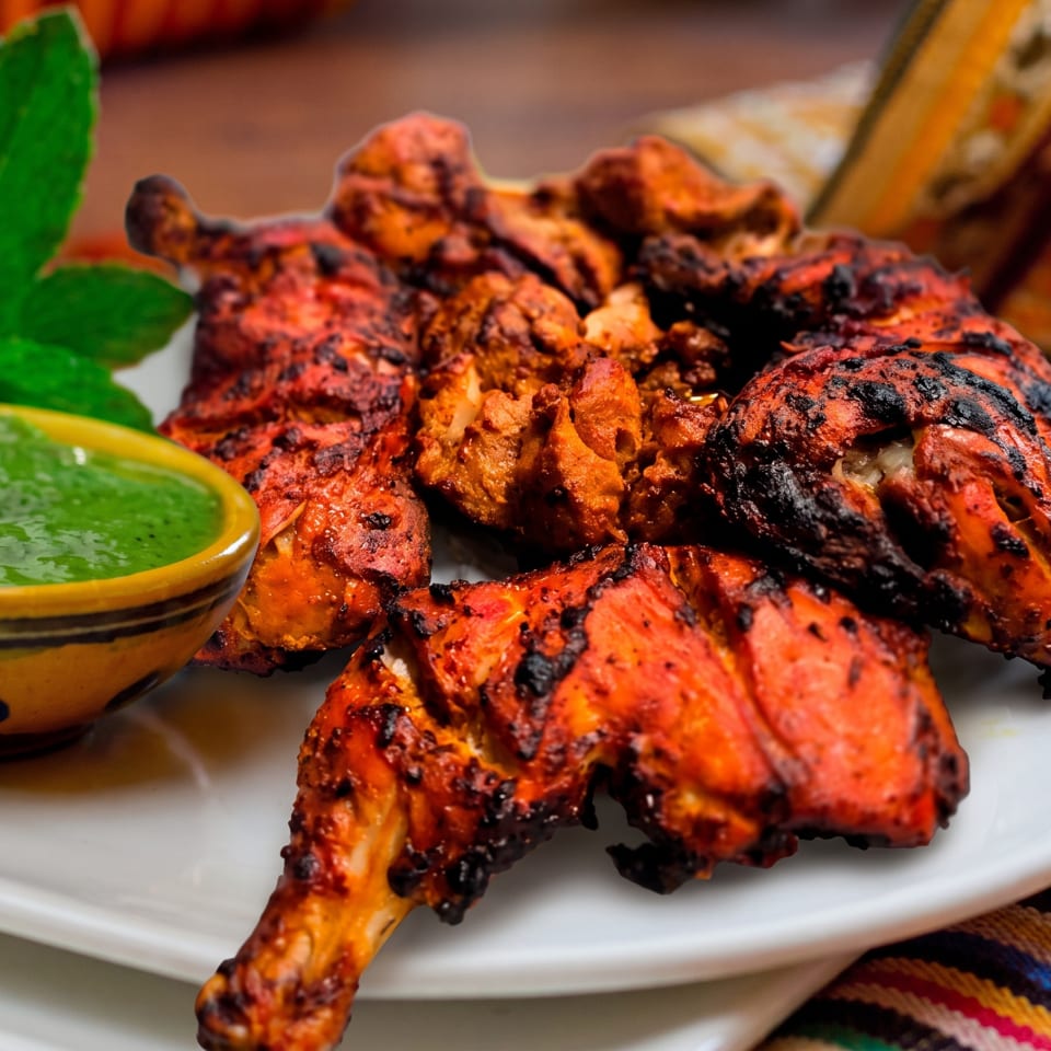 Grilled tandoori chicken pieces with charred edges, served on a white plate with green chutney.